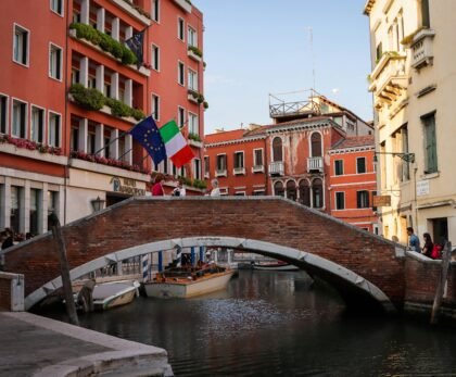 A picturesque view of a brick bridge over a canal in Venice, Italy, with surrounding architecture and vibrant flags.