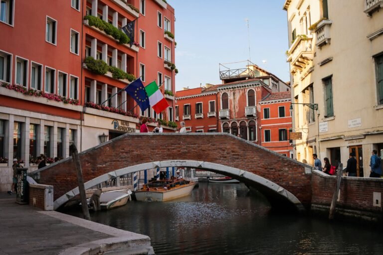 A picturesque view of a brick bridge over a canal in Venice, Italy, with surrounding architecture and vibrant flags.