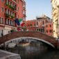 A picturesque view of a brick bridge over a canal in Venice, Italy, with surrounding architecture and vibrant flags.